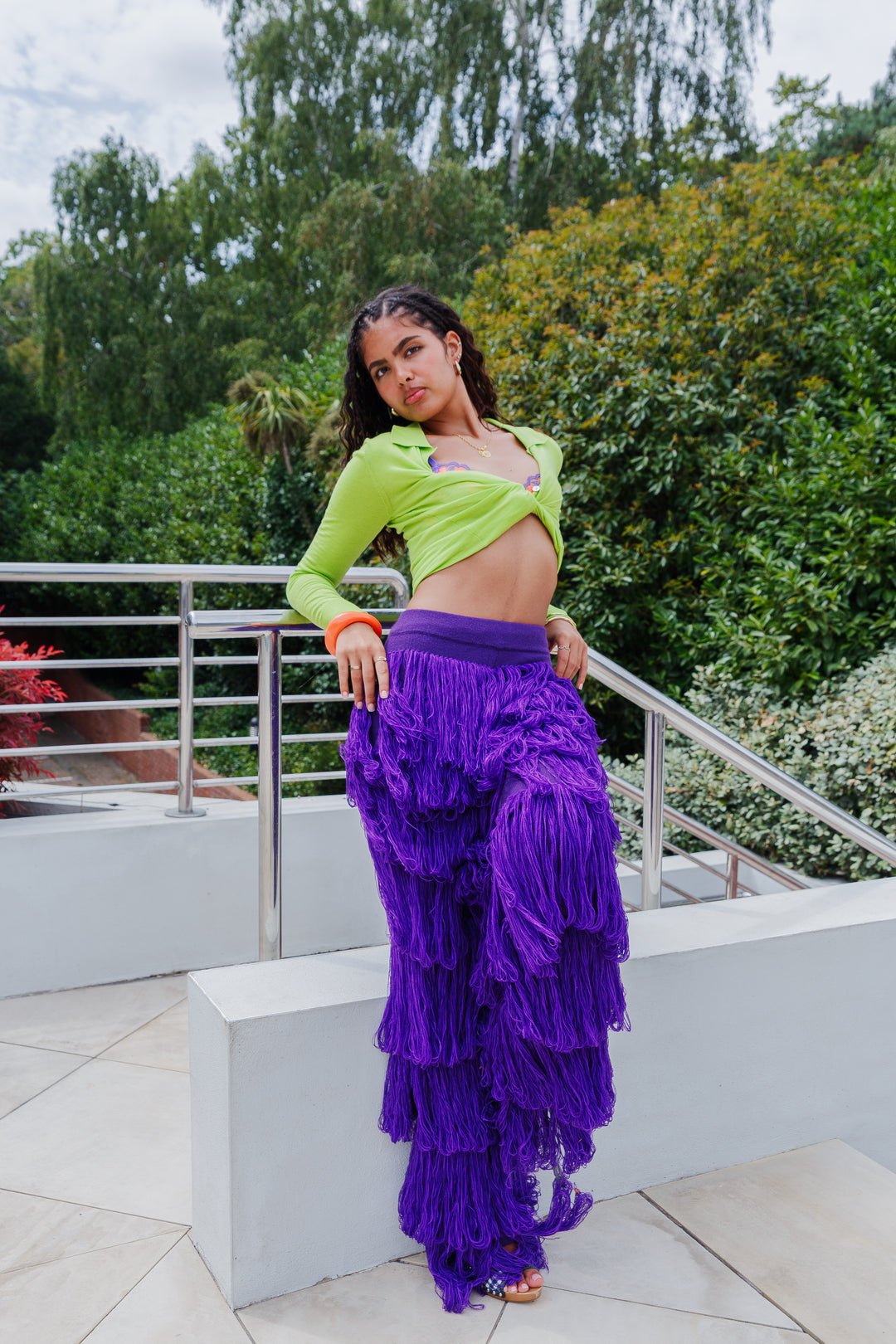 The Archive UK - A model posing in a bright lime green long-sleeve crop top and the Milena Knitted Tassel Trouser Pants in Aubergine purple outdoors.